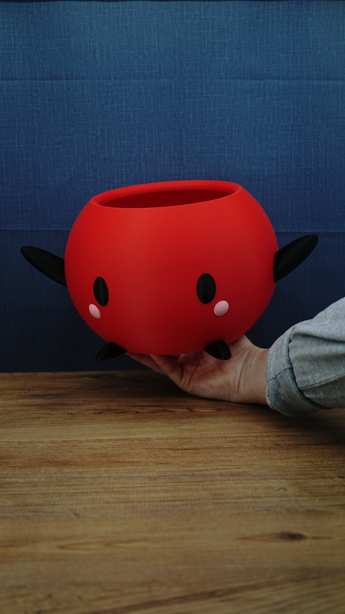 Hand holding a large red Junimo-inspired planter pot with black arms and feet, oval black eyes, and pink cheek details.