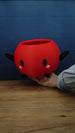 Hand holding a large red Junimo-inspired planter pot with black arms and feet, oval black eyes, and pink cheek details.