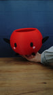 Hand holding a large red Junimo-inspired planter pot with black arms and feet, oval black eyes, and pink cheek details.