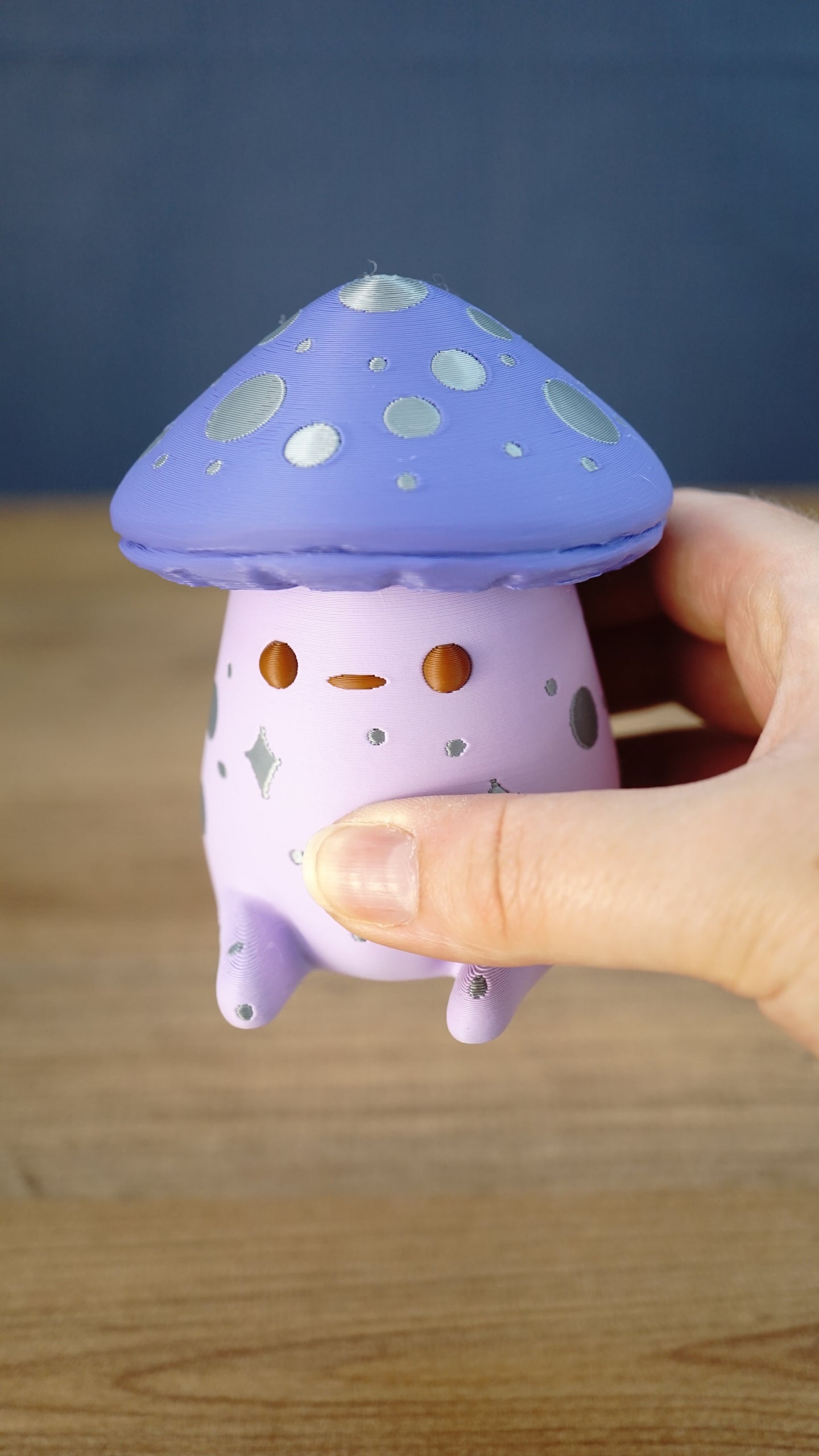 Close-up of a Madrinas Fun Guy figurine being held, showing its lavender cap, silver spots, orange eyes, and pale pink body details.
