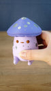 Close-up of a Madrinas Fun Guy figurine being held, showing its lavender cap, silver spots, orange eyes, and pale pink body details.