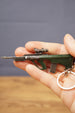 AUG-style metal gun keyring held in hand to show scale, featuring tactical scope, green body, magazine detail and keychain ring