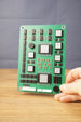 Military circuit board replica held upright by hand, showing green board surface, detailed component layout, silver chips, and white connector pieces.