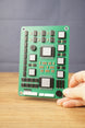Military circuit board replica held upright by hand, showing green board surface, detailed component layout, silver chips, and white connector pieces.