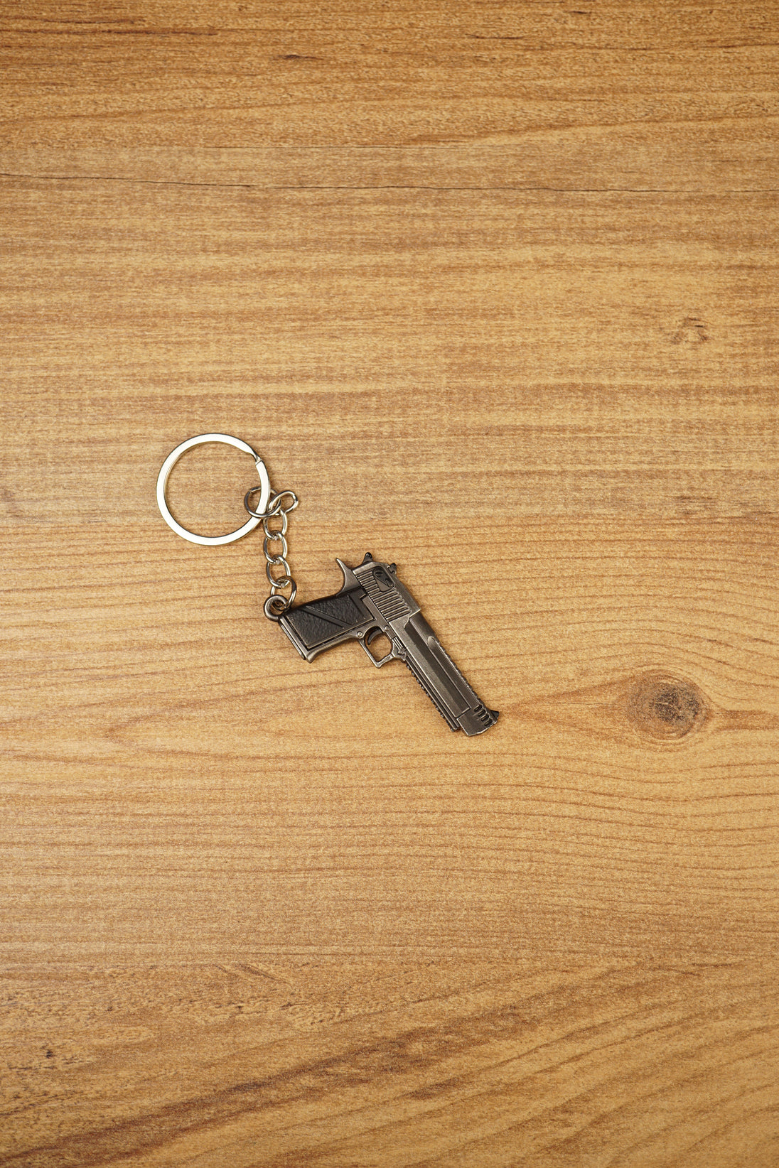 Miniature Deagle pistol keychain displayed on a wooden tabletop with black grip detail, gunmetal body and attached keyring chain