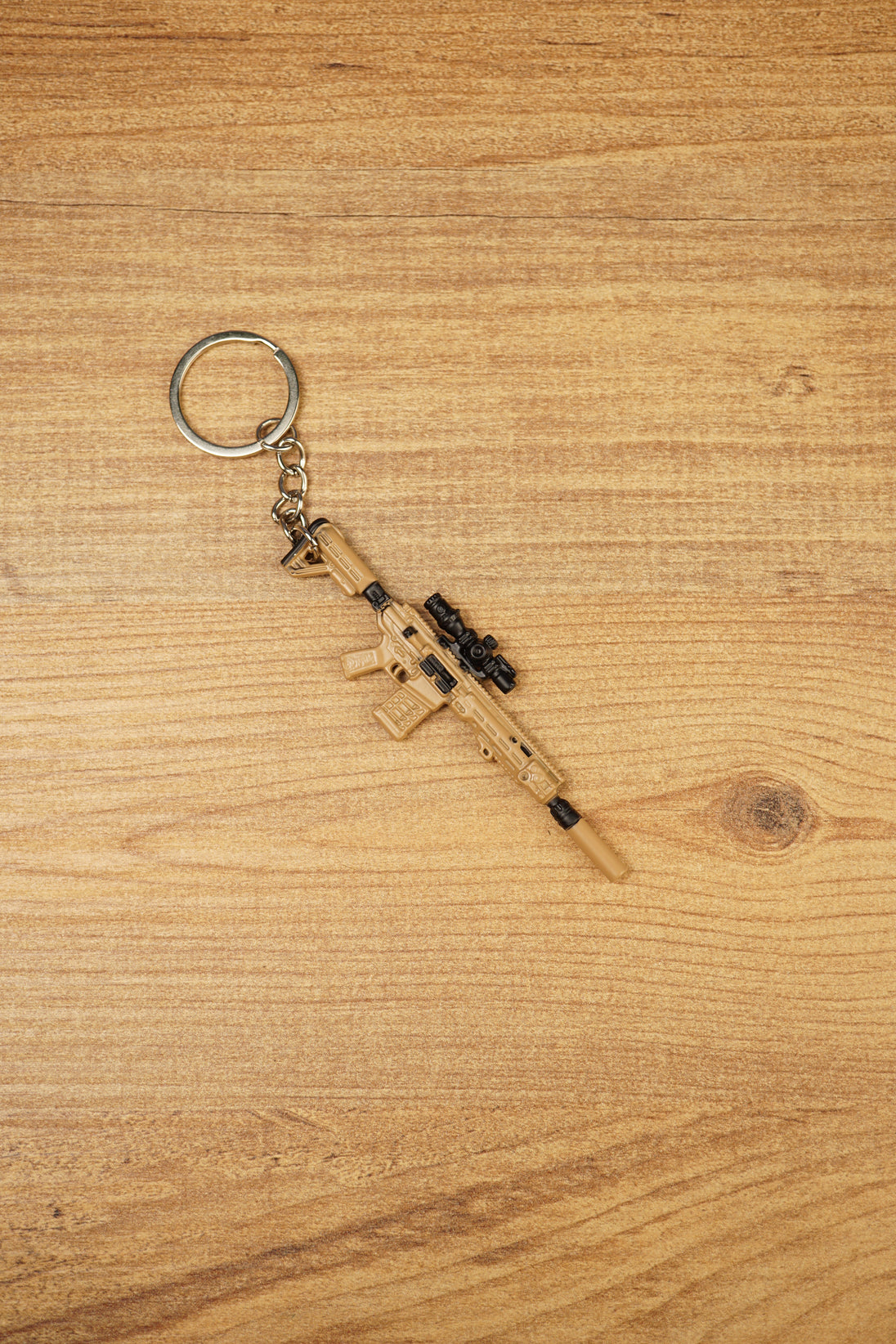 Mini tactical rifle keychain with tan body, black scope and metal keyring, shown diagonally on wood for FPS gamer accessory listing