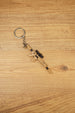 Mini tactical rifle keychain with tan body, black scope and metal keyring, shown diagonally on wood for FPS gamer accessory listing