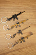 Angled display of four tactical rifle keychains on a wooden background showing different FPS gamer accessory styles.