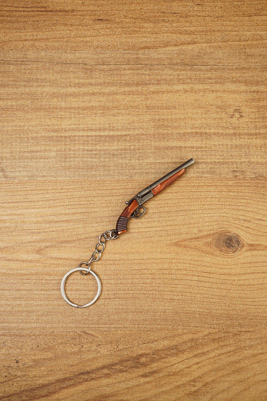 Mini double-barrel shotgun keychain displayed diagonally on wood, featuring gunmetal barrels, brown stock detail and silver keyring