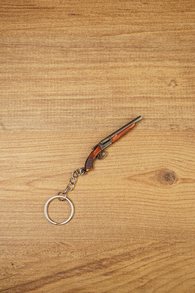 Mini double-barrel shotgun keychain displayed diagonally on wood, featuring gunmetal barrels, brown stock detail and silver keyring