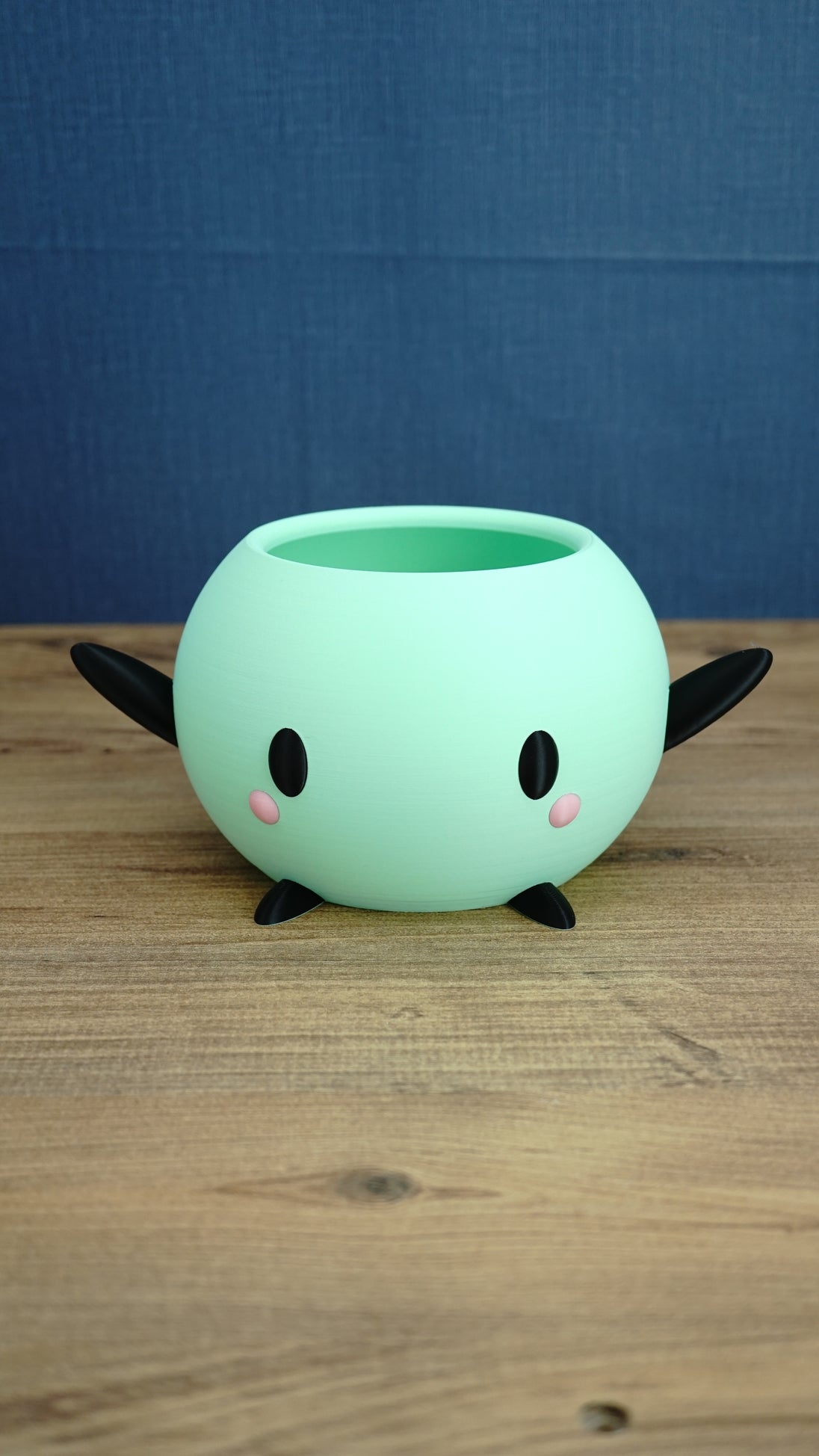 Mint green Stardew Valley Junimo planter pot with black arms, black feet, oval eyes, and pink cheeks on a wooden tabletop.