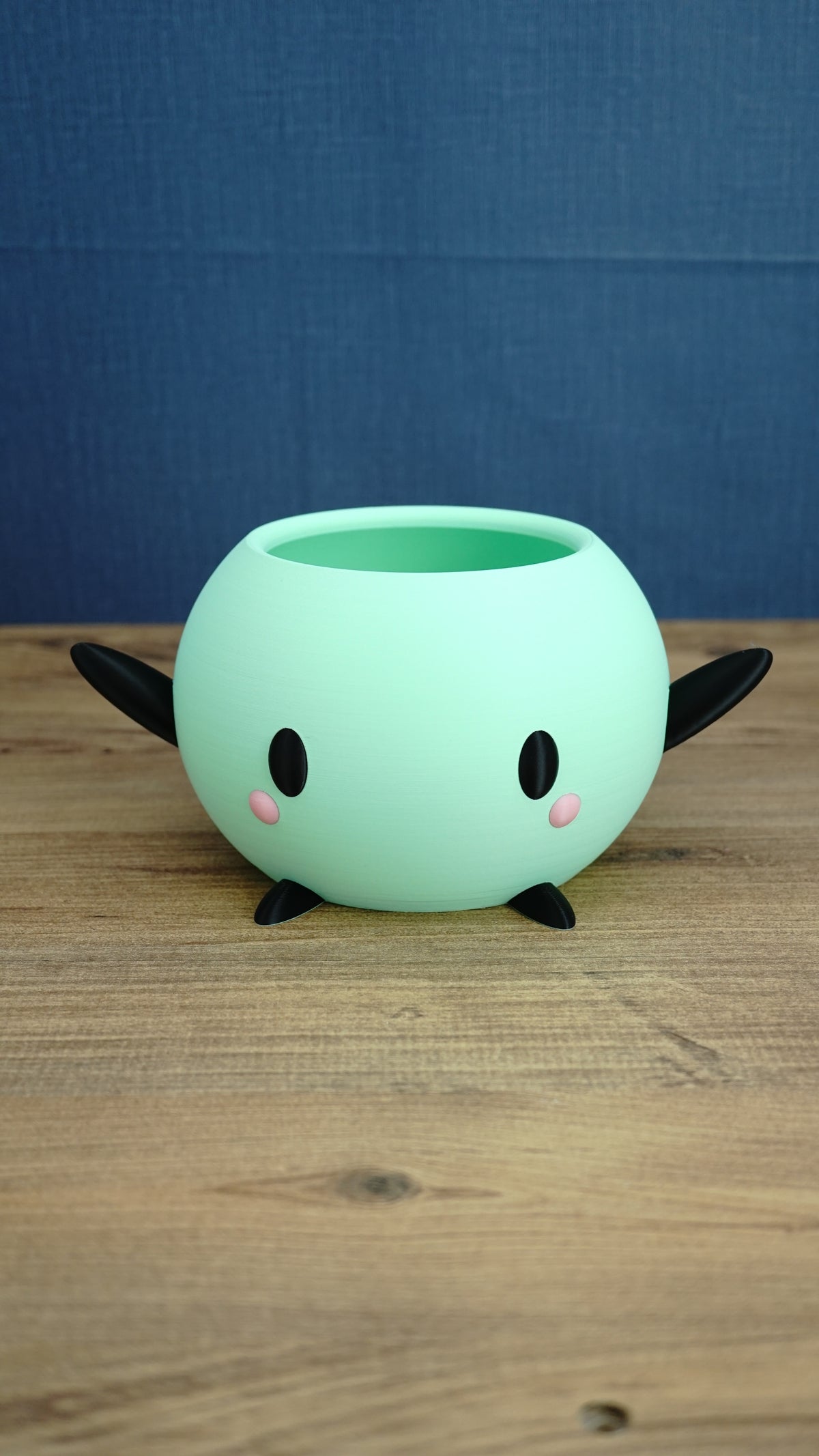 Mint green Stardew Valley Junimo planter pot with black arms, black feet, oval eyes, and pink cheeks on a wooden tabletop.