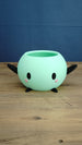 Mint green Stardew Valley Junimo planter pot with black arms, black feet, oval eyes, and pink cheeks on a wooden tabletop.