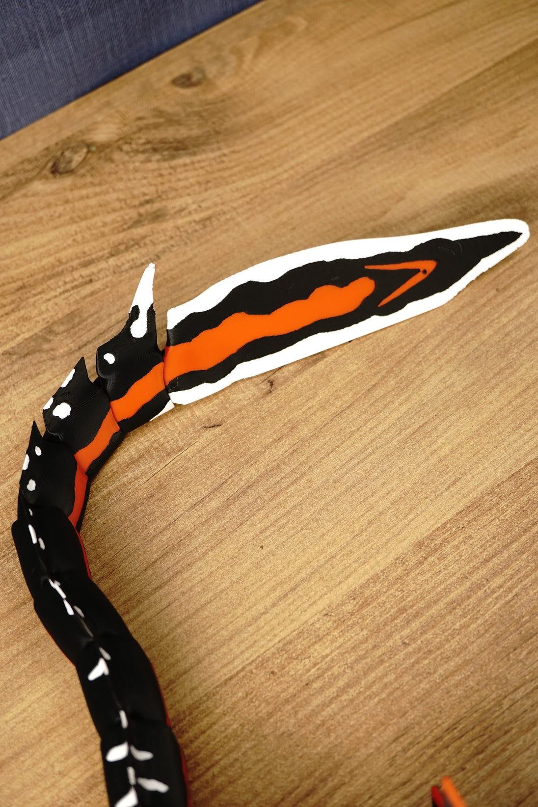 Needler Mango model tail displayed on a wooden surface, showing the articulated black body segments and large black, orange and white tail fin.
