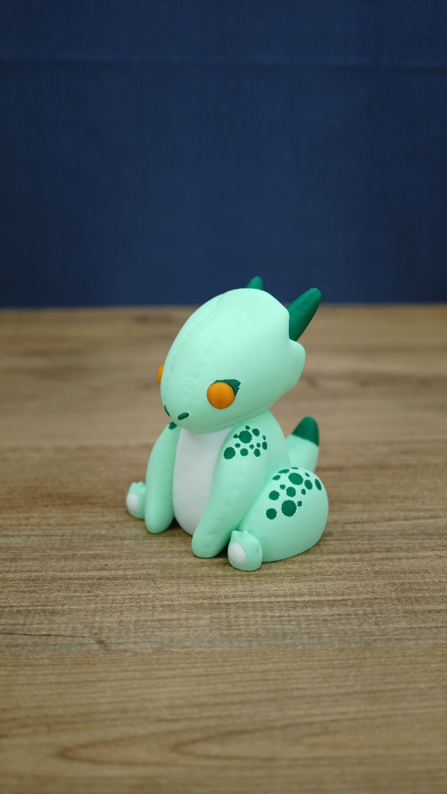 Side angle of Nibbles the Drake inspired Palia figure showing the cute seated baby drake pose, green spotted legs, horns, tail and white paw details