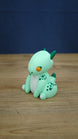 Side angle of Nibbles the Drake inspired Palia figure showing the cute seated baby drake pose, green spotted legs, horns, tail and white paw details