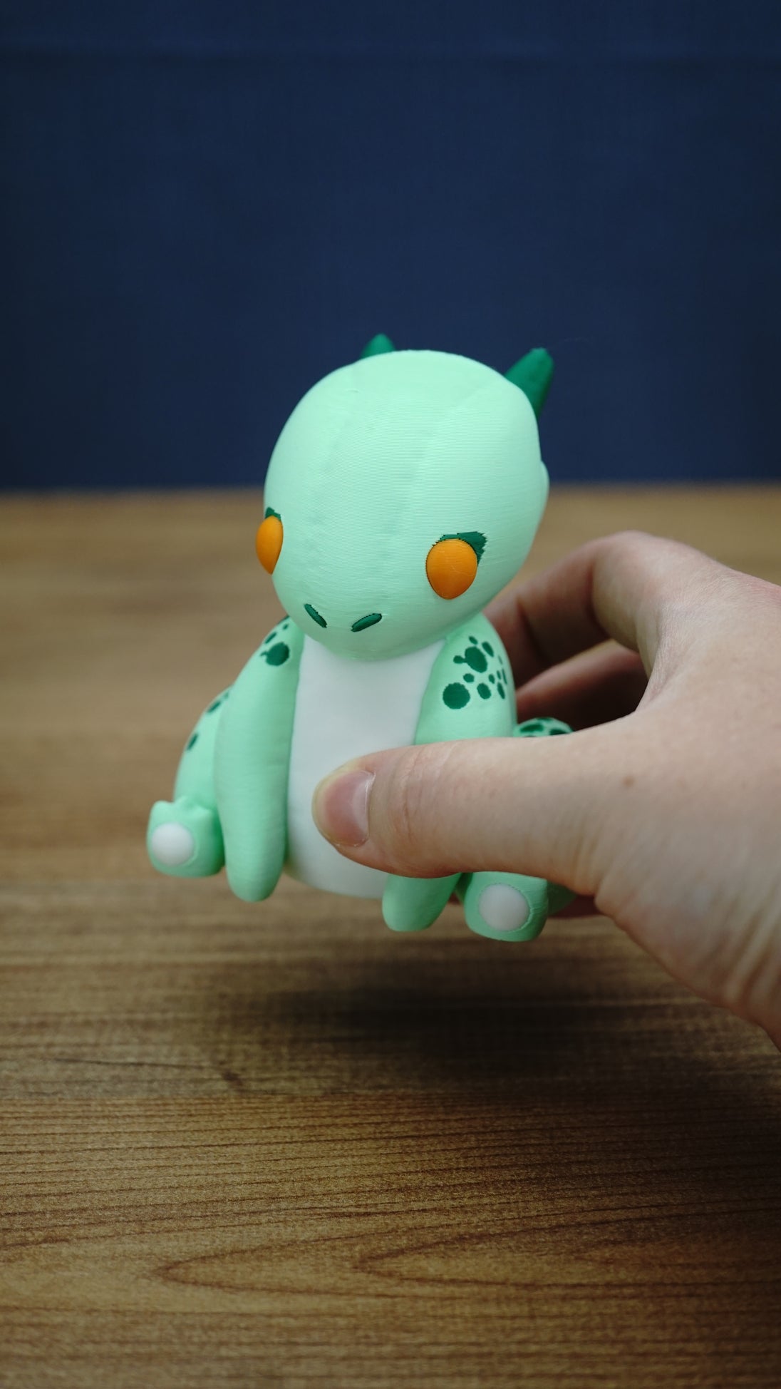 Nibbles the Drake Palia inspired figure held in a hand to demonstrate its compact collectible size, with orange eyes, green horns and white belly visible