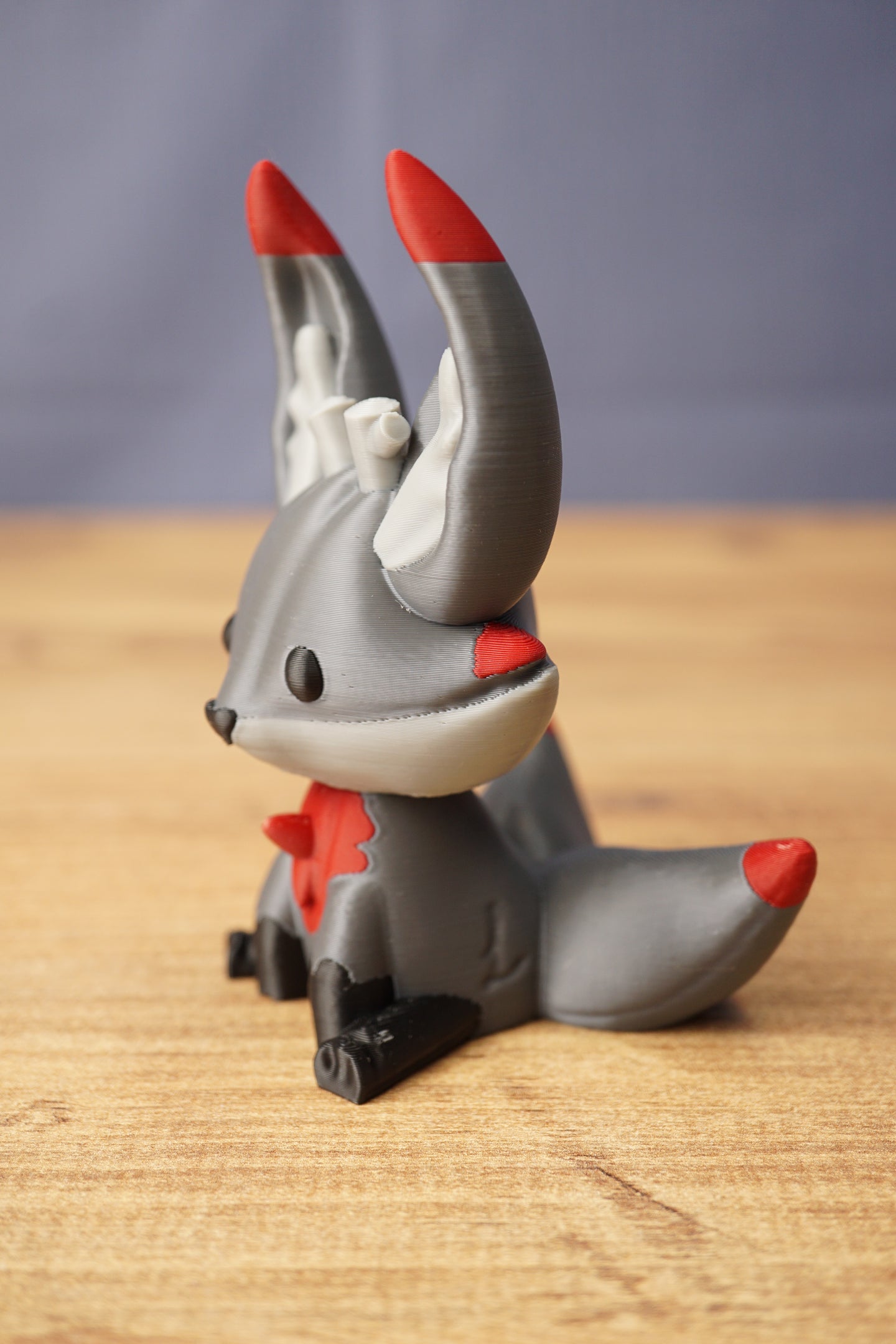 Obsidian Kitsuu fox figure shown from the side with grey body, red ear tips, white inner ears and curled tail on wooden surface