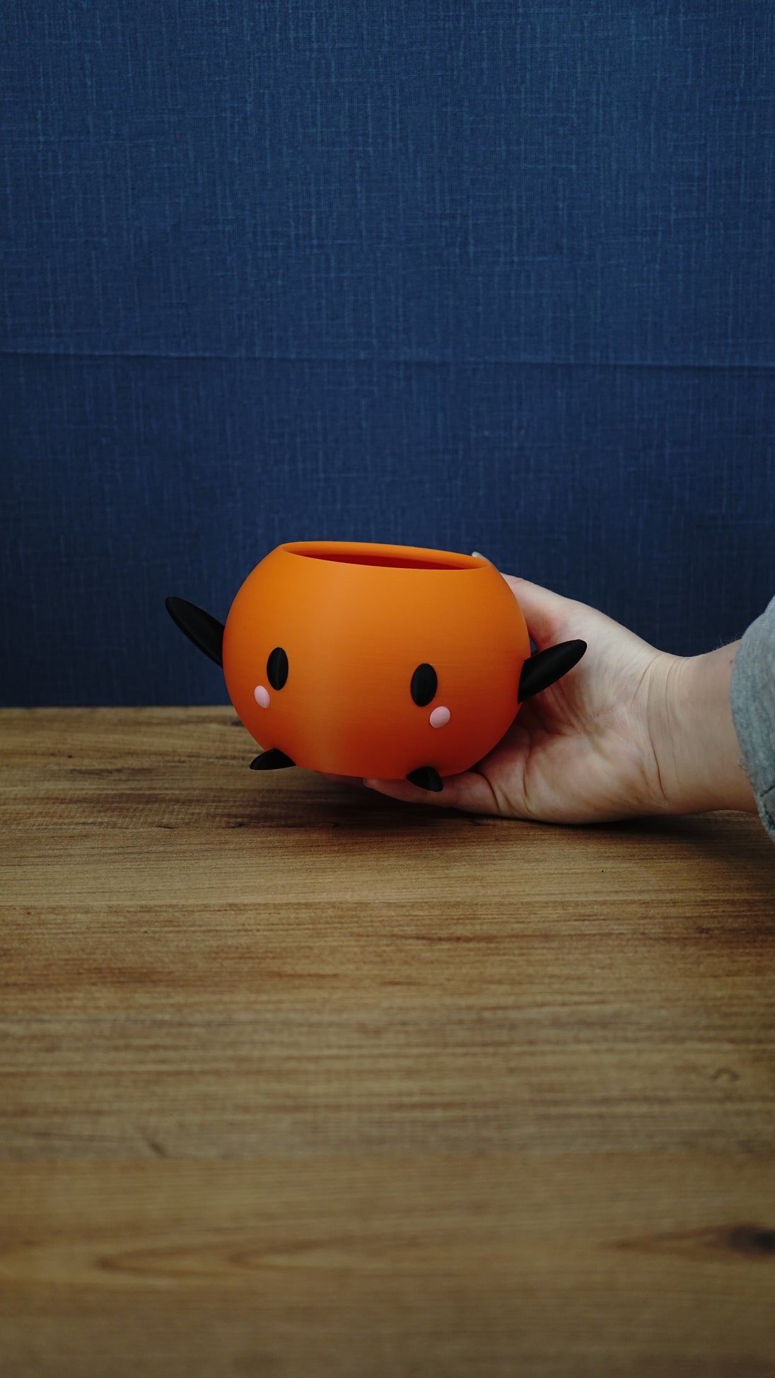 Hand holding a small orange Junimo-inspired planter pot with black arms and feet, oval black eyes, and pink cheek details.