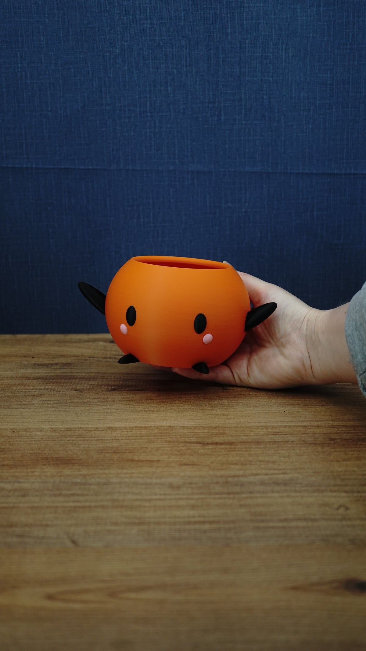 Hand holding a small orange Junimo-inspired planter pot with black arms and feet, oval black eyes, and pink cheek details.