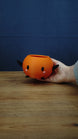 Hand holding a small orange Junimo-inspired planter pot with black arms and feet, oval black eyes, and pink cheek details.