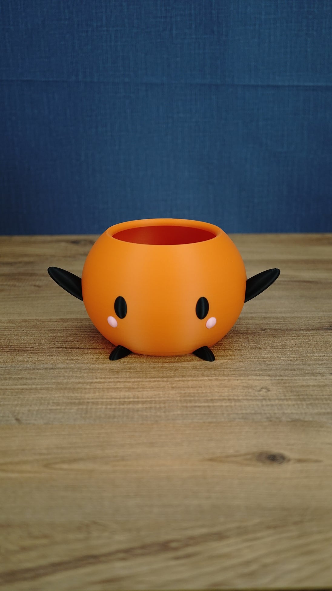 Orange Stardew Valley Junimo planter pot with black arms, black feet, oval black eyes, and pink cheeks against a blue backdrop.