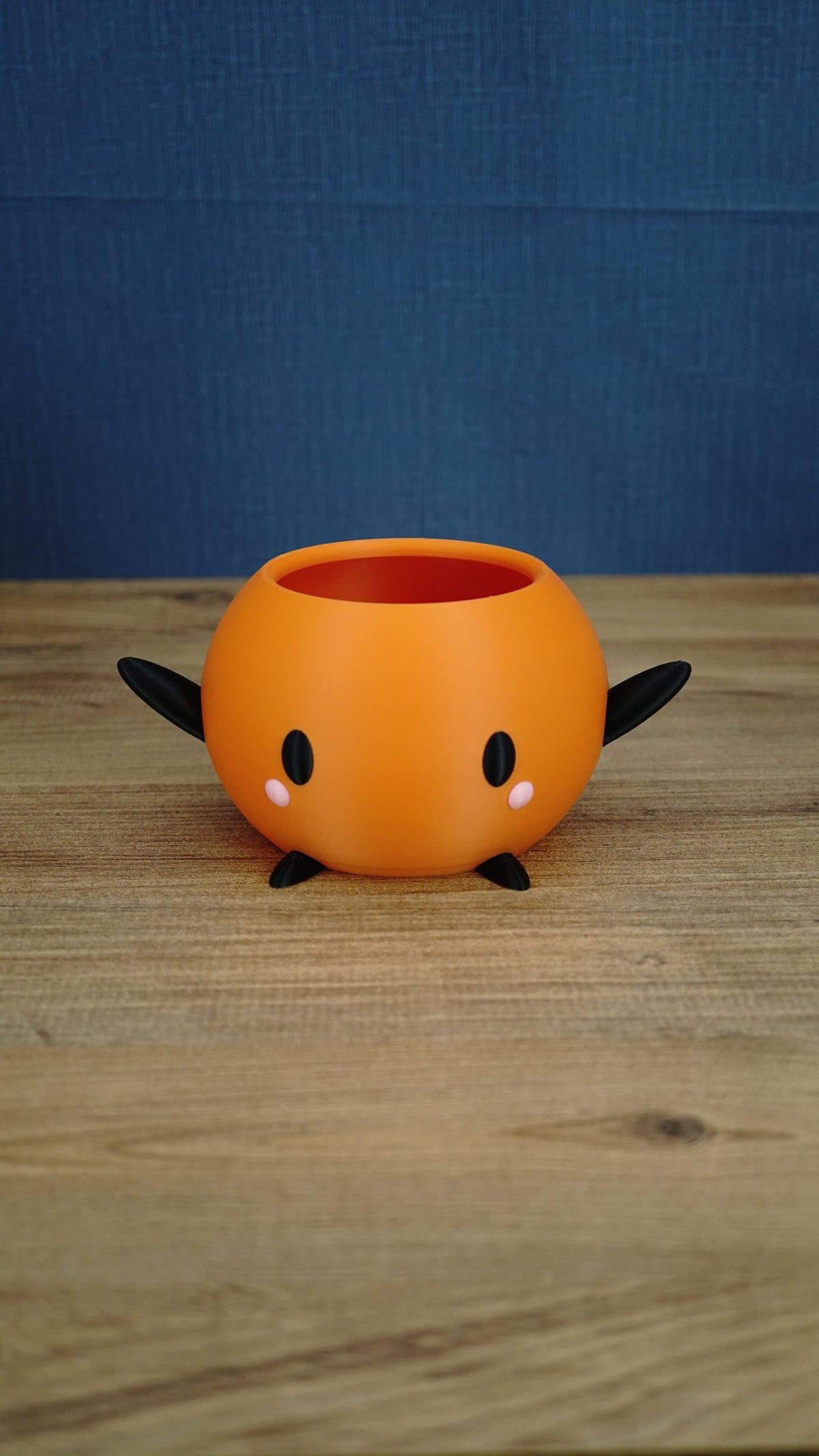 Orange Stardew Valley Junimo planter pot with black arms, black feet, oval black eyes, and pink cheeks against a blue backdrop.