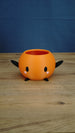 Orange Stardew Valley Junimo planter pot with black arms, black feet, oval black eyes, and pink cheeks against a blue backdrop.