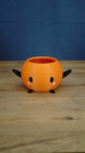 Orange Stardew Valley Junimo planter pot with black arms, black feet, oval black eyes, and pink cheeks against a blue backdrop.