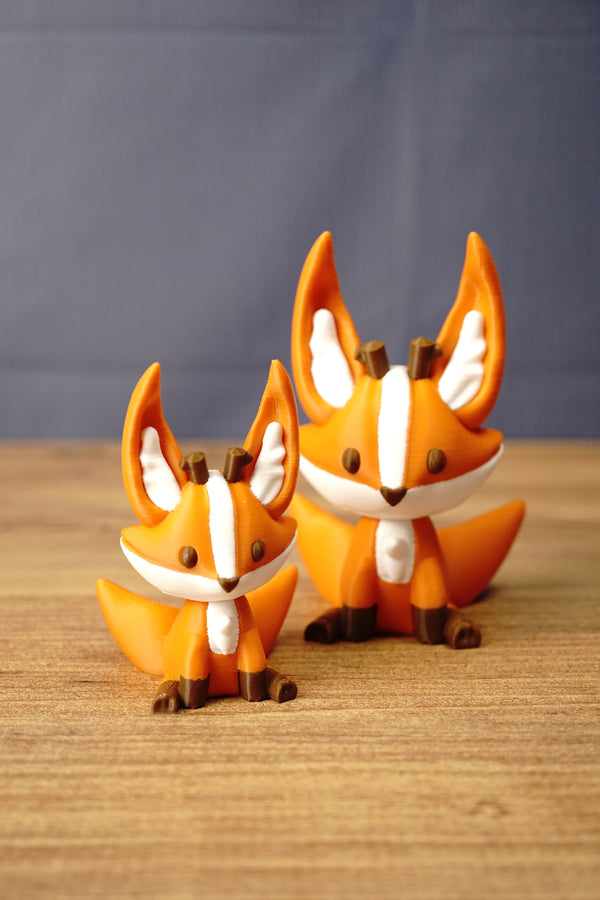 Small and large orange Ancient Kitsuu-inspired 3D-printed fox figures displayed together on a wooden tabletop.