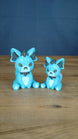 Two blue 3D printed Palcat plush figures with dark grey ear tufts, amber eyes, white paw pads, and white chest details on a wooden tabletop.