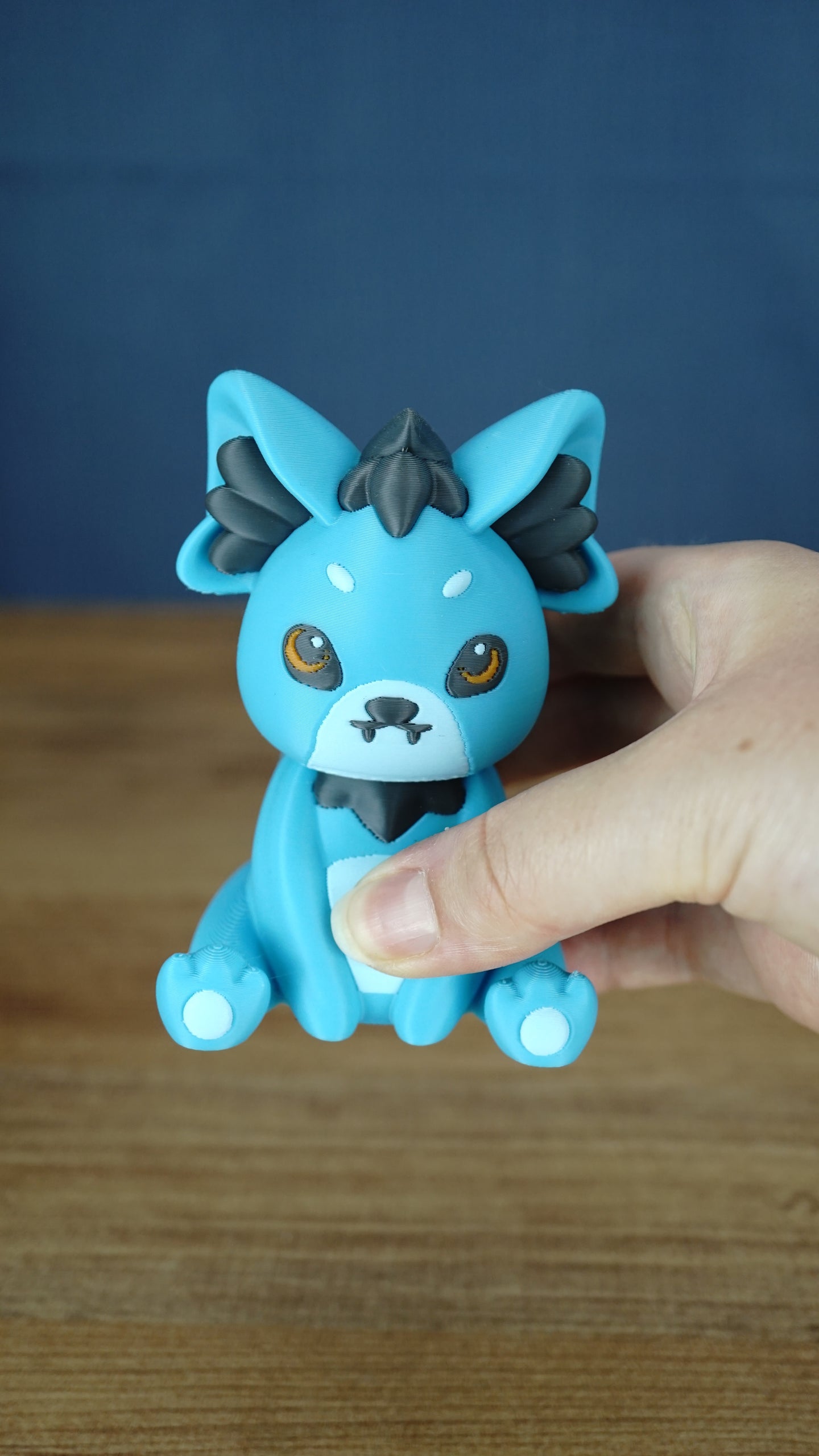 Handheld close-up of a blue 3D printed Palcat plush figure with amber eyes, dark grey ear and chest accents, white muzzle, tiny fangs, and white paw pads.
