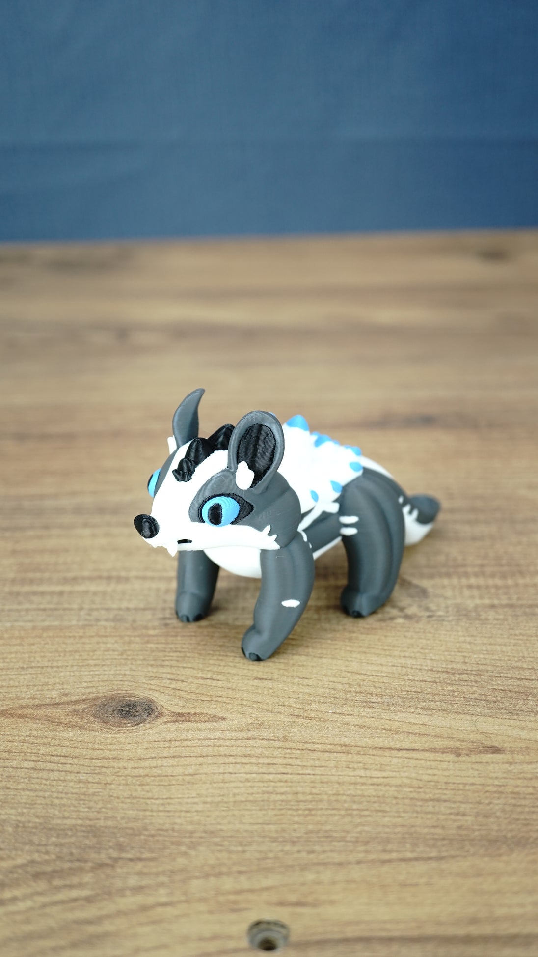 Bluebristle Muujin plush-inspired figure from Palia shown at an angle, featuring bright blue eyes, a grey body, a white mane and turquoise blue spikes.