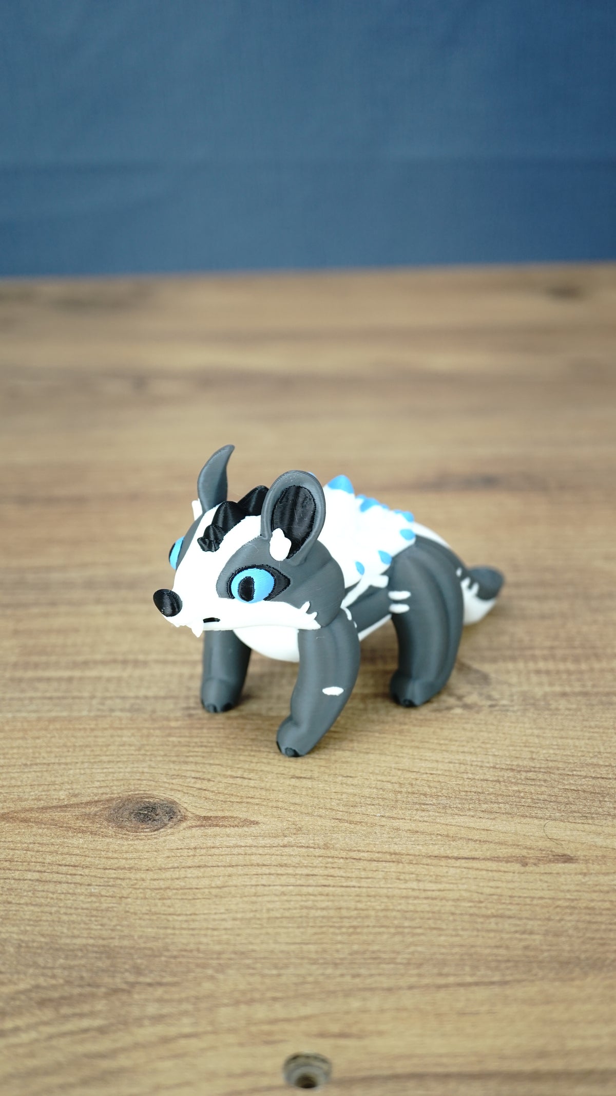 Bluebristle Muujin plush-inspired figure from Palia shown at an angle, featuring bright blue eyes, a grey body, a white mane and turquoise blue spikes.