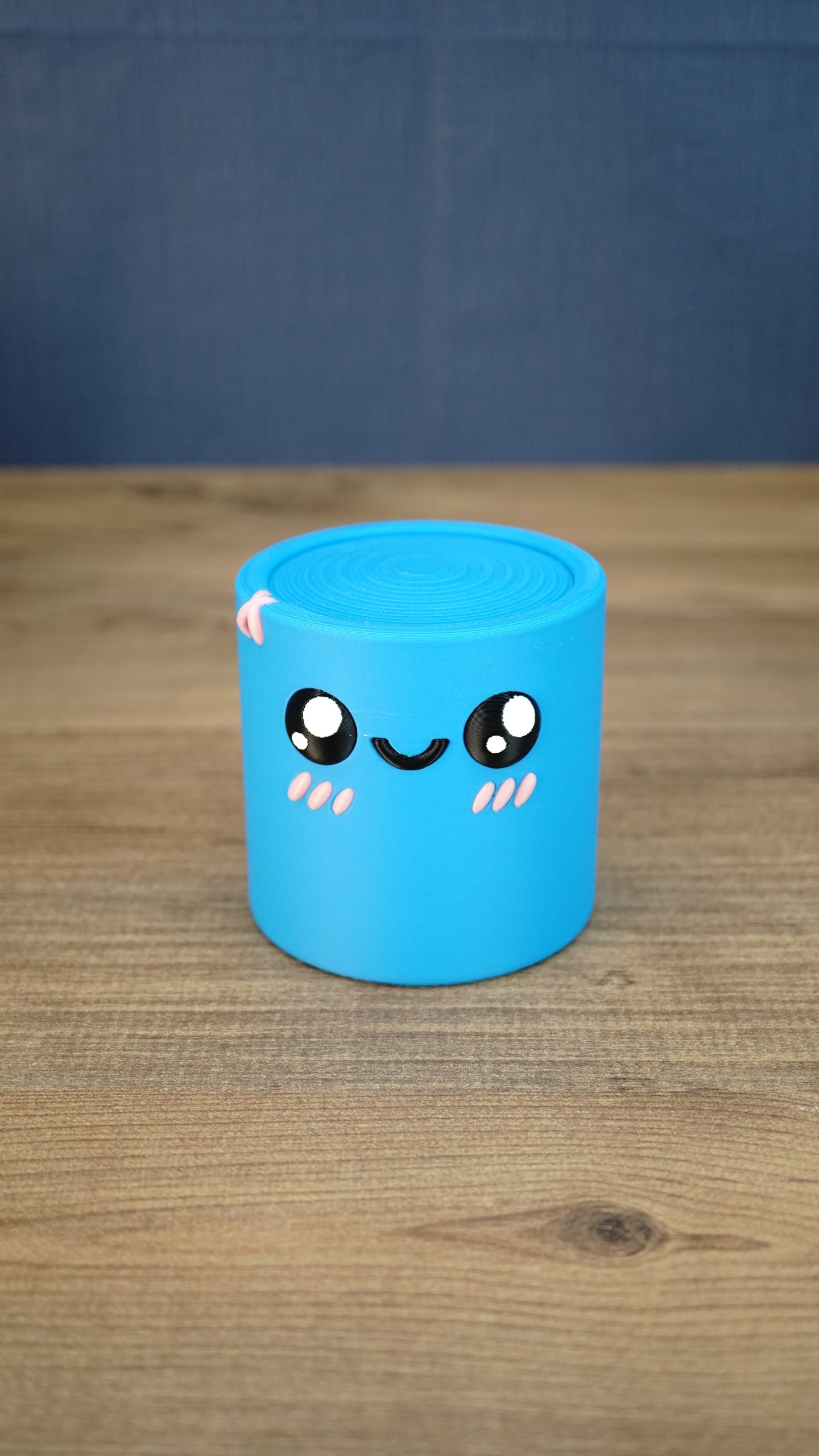 Front view of the blue Palia Cyl Cylinder Collectible with black eyes, a smiling face, and pink cheeks.