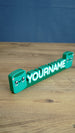 Angled view of custom Palia Cyl-inspired gamer nameplate with raised white lettering and cute green characters for desk or gaming room decor