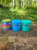 Three Palia Cyl Cylinder Collectible figures in rainbow, blue, and teal displayed on a tree stump in a forest setting.