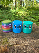 Three Palia Cyl Cylinder Collectible figures in rainbow, blue, and teal displayed on a tree stump in a forest setting.