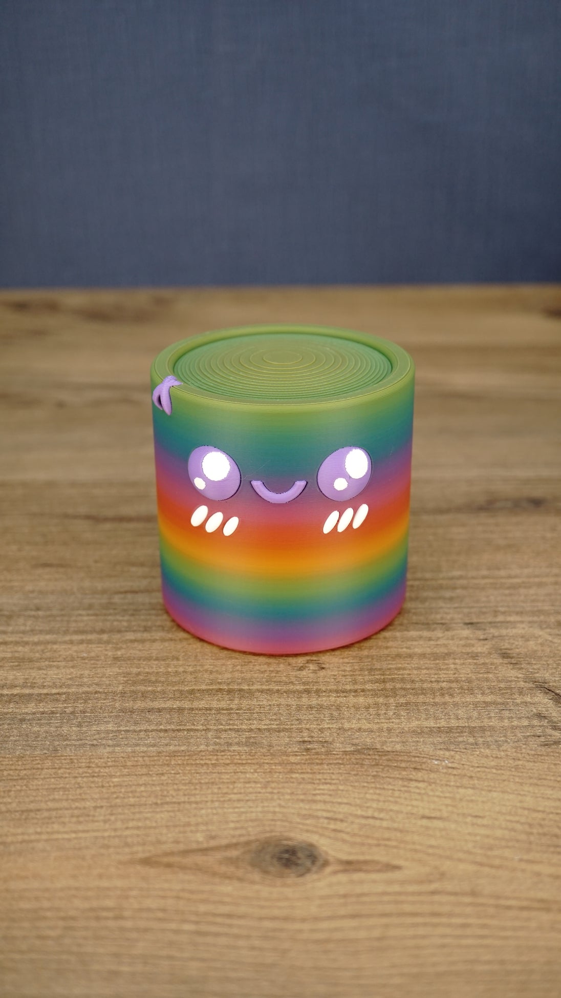 Front view of a rainbow Palia Cyl Cylinder collectible with purple eyes, a smiling face, and white cheek details.
