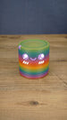 Front view of a rainbow Palia Cyl Cylinder collectible with purple eyes, a smiling face, and white cheek details.