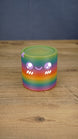 Front view of a rainbow Palia Cyl Cylinder collectible with purple eyes, a smiling face, and white cheek details.