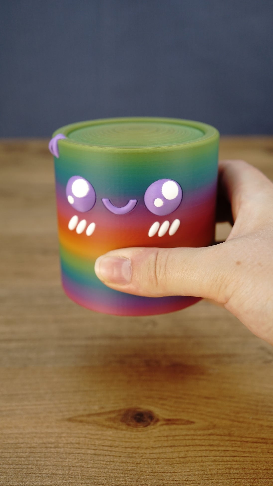 Close-up of a rainbow Palia Cyl Cylinder collectible being held, showing its purple eyes, smiling face, cheek details, and 3D printed texture.