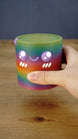 Close-up of a rainbow Palia Cyl Cylinder collectible being held, showing its purple eyes, smiling face, cheek details, and 3D printed texture.