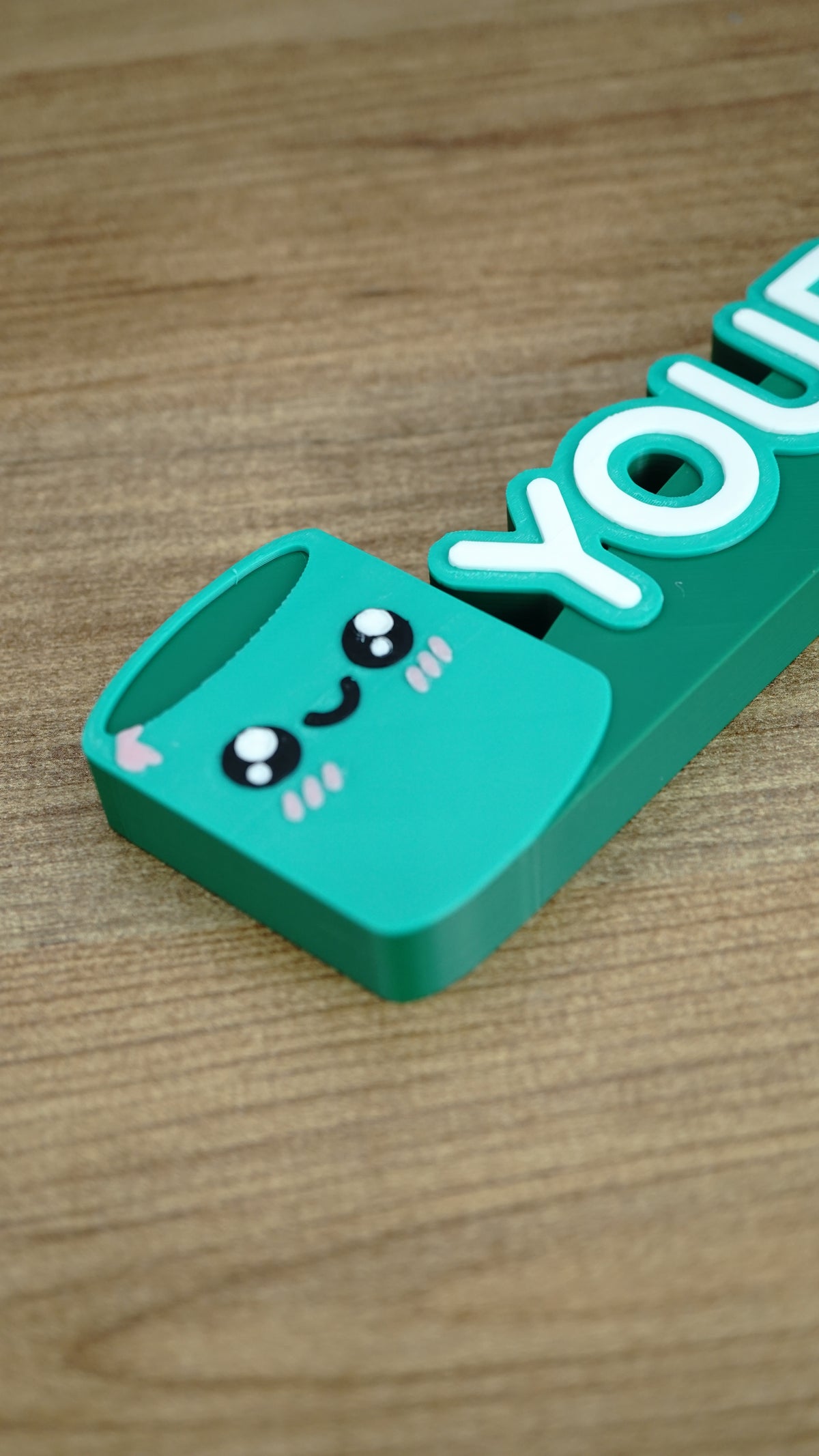 Close-up of cute green Cyl-inspired character beside raised personalized lettering on a custom Palia gamer nameplate for desks and shelves