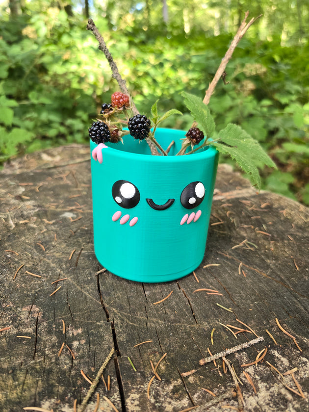Teal Palia Cyl Pen Pot with a cute smiling face, holding blackberry branches on a tree stump in a forest.
