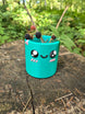 Teal Palia Cyl Pen Pot with a cute smiling face, holding blackberry branches on a tree stump in a forest.