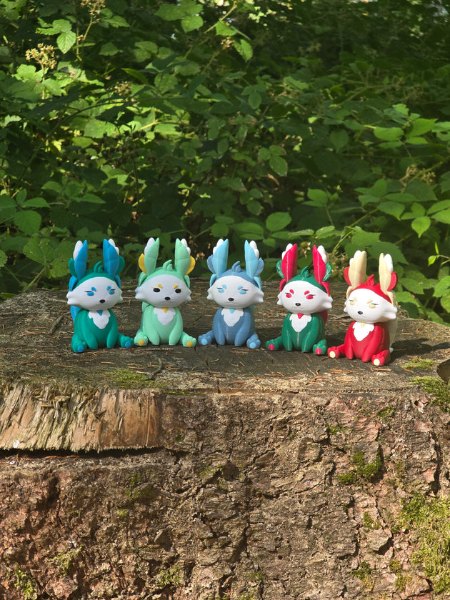 Five colourful 3D printed Palia Flutterfox plush figures displayed in a row on a tree stump in a forest.