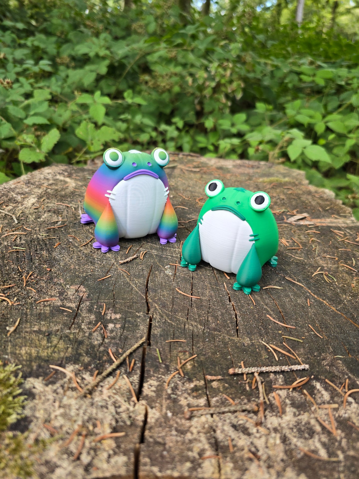 Green and rainbow Frogbert 3D printed plush figures displayed on a tree stump in a forest.