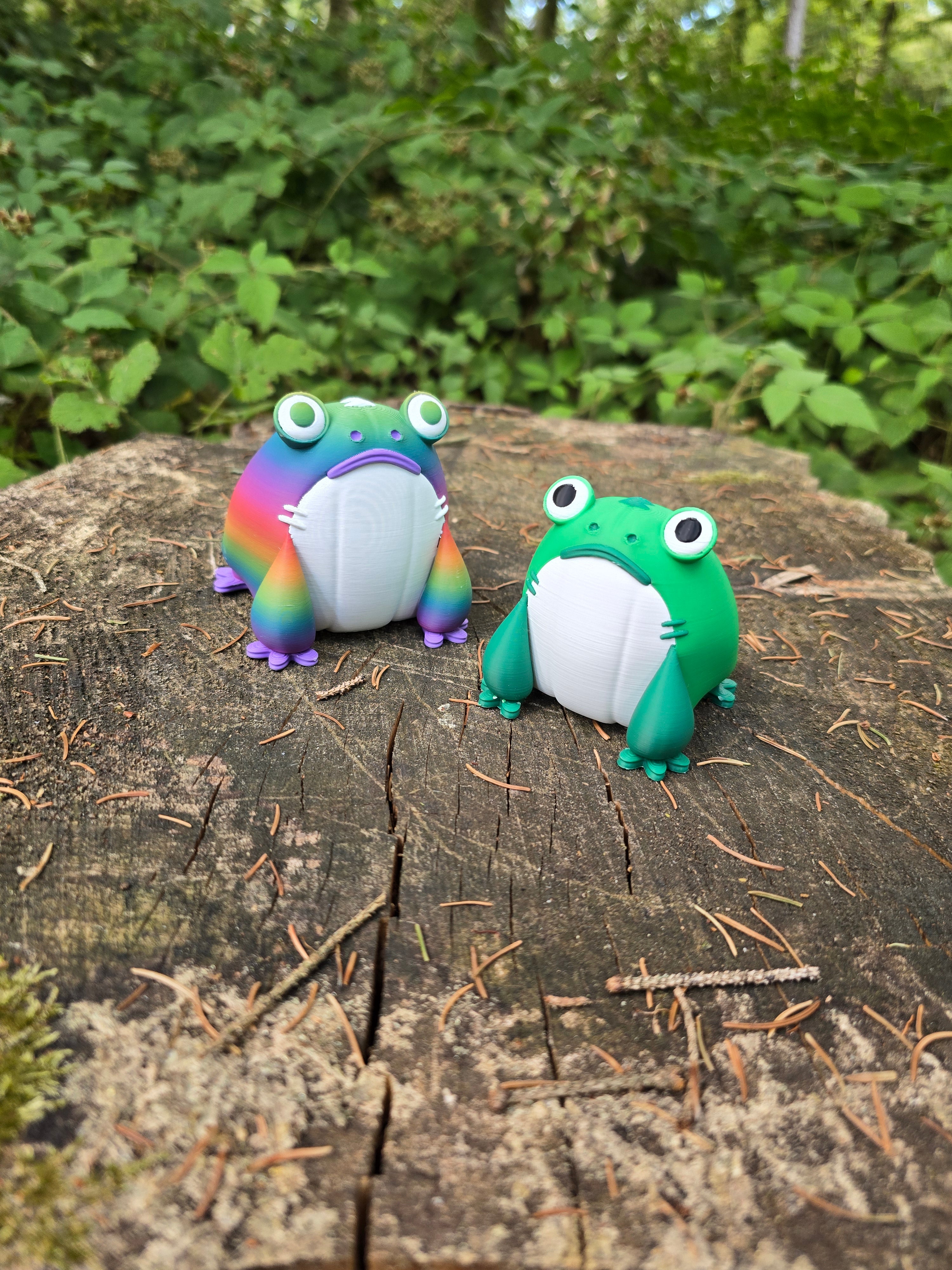 Green and rainbow Frogbert 3D printed plush figures displayed on a tree stump in a forest.