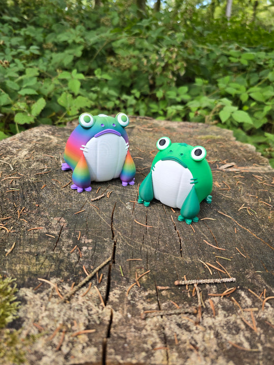 Green and rainbow Frogbert 3D printed plush figures displayed on a tree stump in a forest.